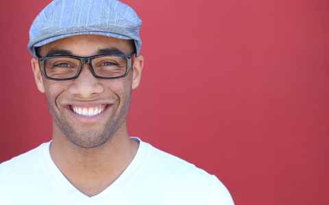 man with newsboy cap, trendy black glasses, and white v neck shirt smiling at the camera