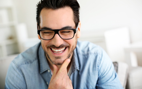 man with black glasses, holding his chin and smiling 